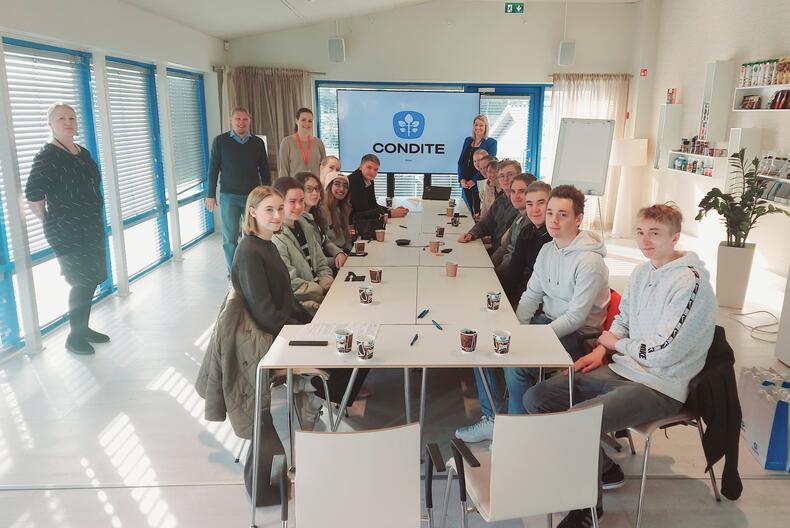 Opiskelijat istuvat neuvottelupöydän ääressä Conditen vaaleissa tiloissa, opettajia ja henkilökuntaa seisoo pöydän ympärillä.