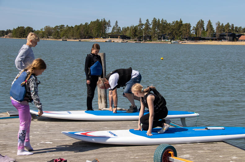 Laiturilla mies neuvoo sup-laudan päällä, miten laudalla ollaan. Lapset seuraavat vieressä.