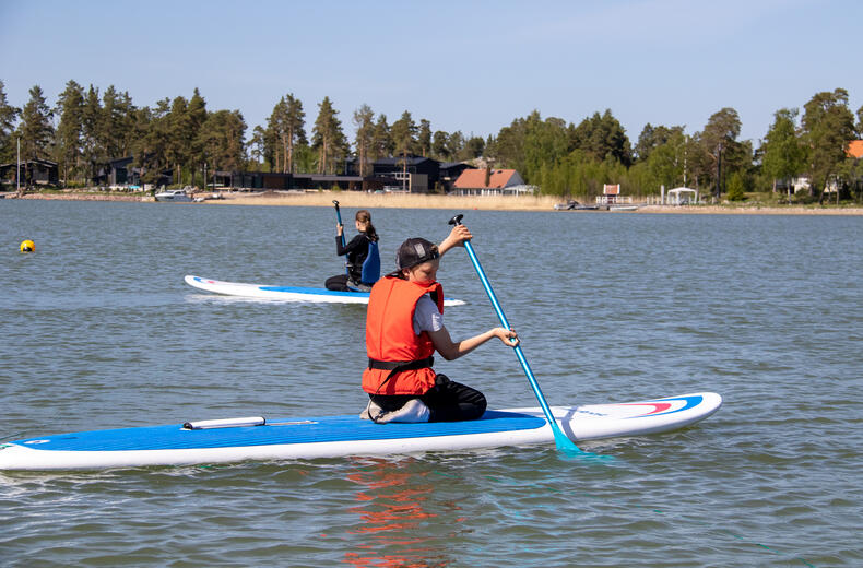 Kaksi koululaista istuu sup-laudoilla polviltaan ja meloo eteenpäin.
