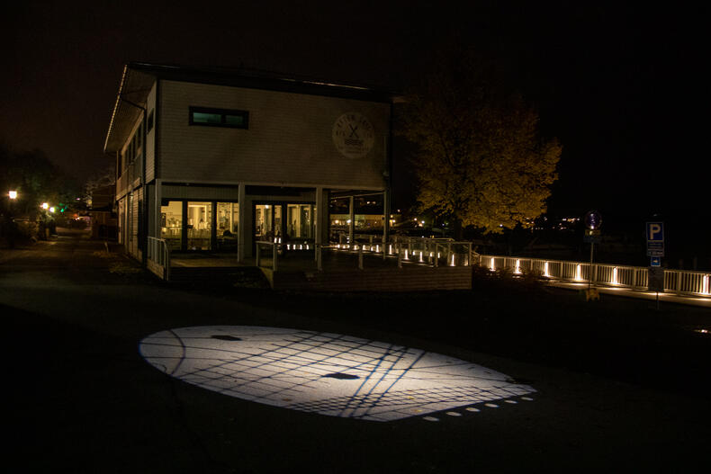 Purjeveneiden mastoja ja köysistöä heijastuu asfaltille, taustalla rannan ravintola ja valaistua laituria.