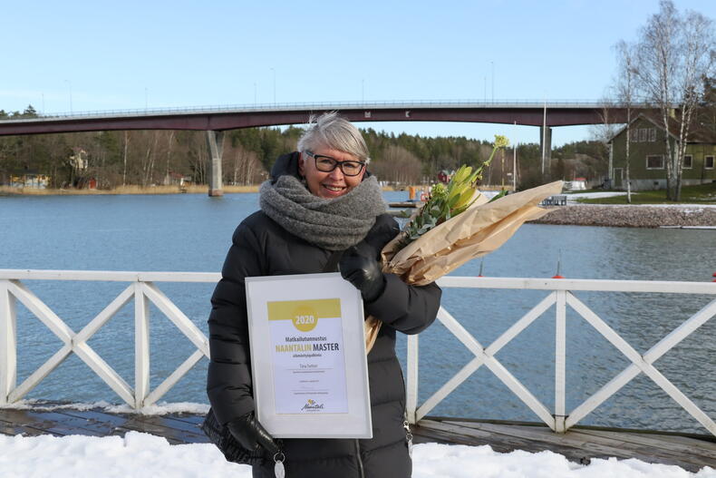 Hymyilevä Tiina Tunturi käsissään tunnustus ja palkintokukat, taustalla meri.