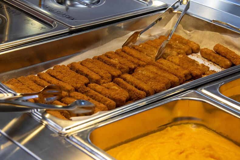 Kouluruokalinjastolla on kalapuikkoja, molemmin puolin linjastoa on ottimet ruokaan. Kalapuikkojen vieressä on oranssia muusia.