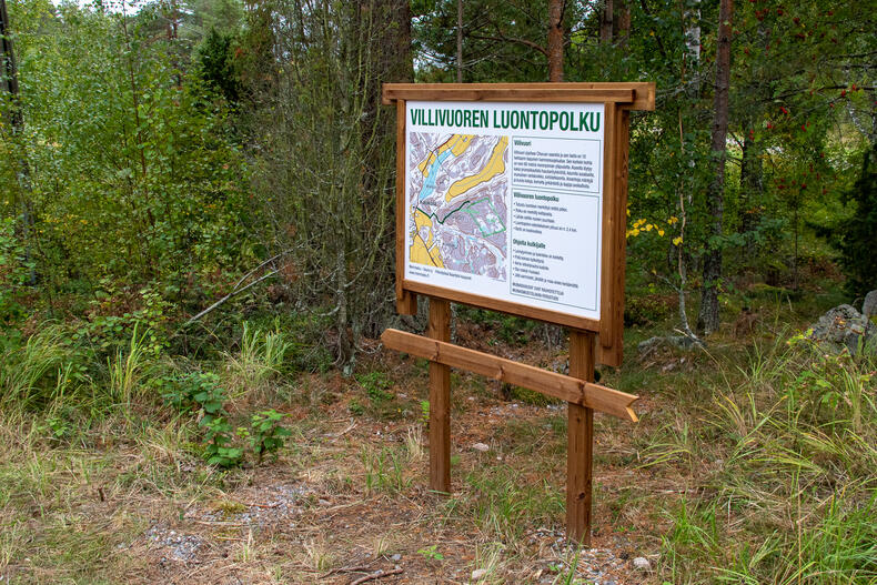 Metsän reunassa on kyltti, jossa lukee Villivuoren luontopolku ja kartta reitistä sekä tekstiä.