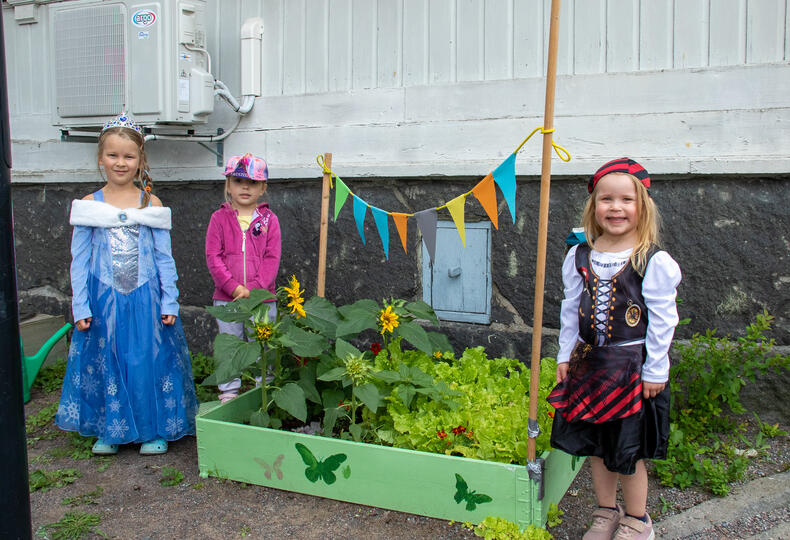 Kolme lasta seisoo hymyillen lavakauluksen ympärillä, siinä on pieni kasvimaa. Yhdellä lapsista on Frozen-prinsessamekko, toisella merirosvoasu.