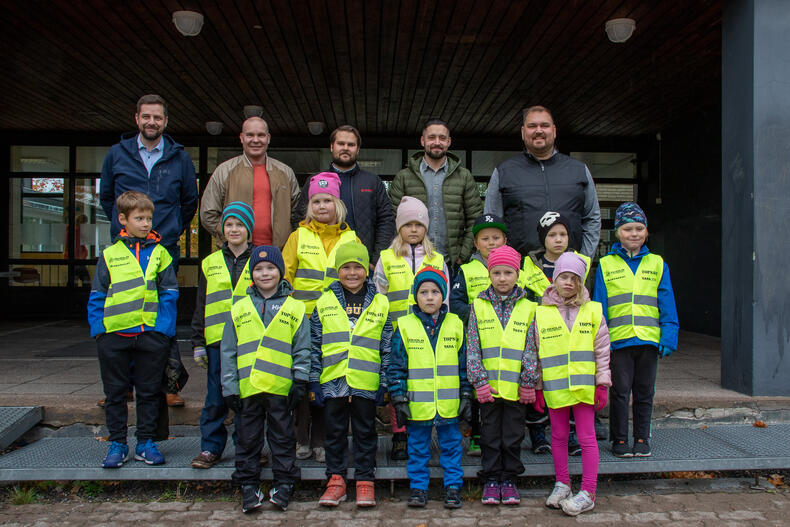 Koulurakennuksen edessä seisoo kolmessa rivissä aikuisia ja lapsia: Edessä kahdessa rivissä on keltaisiin heijastinliiveihin pukeutuneita ekaluokkalaisia, ja heidän takanaan viisi miestä.