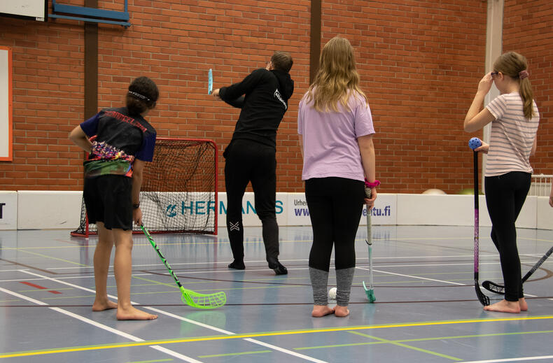 Eemeli Salin laukaisee sählymailalla pallon maaliin, lapsia seisoo rivissä hänen takanaan katsomassa.