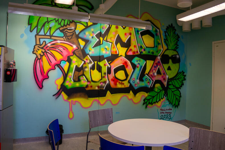 Sisätilan seinälle maalattu graffiti, jossa tyylitellyt ananas, palmu, aurinkovarjo sekä teksti "Emoluola".