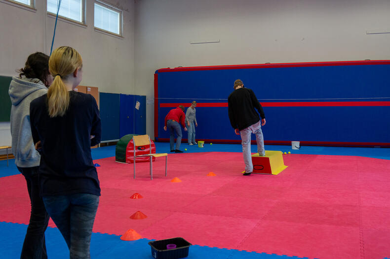 Liikuntasalissa nuoria kulkemassa lasit päässä erilaisten esteiden yli ja heittelemässä palloja, toiset nuoret odottavat vuoroaan.