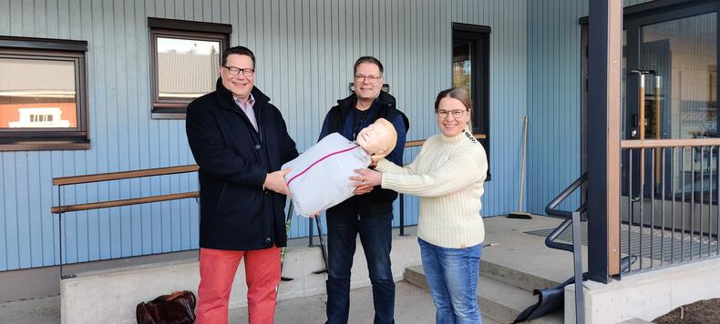 Petri Parviainen, Kimmo Kuusimäki ja Velkuan koulunjohtaja Päivi Soukka pitelevät elvytysnukkea yhdessä keskellään.