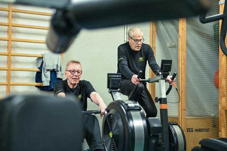 Kaksi miestä treenaa kuntosalilla. Toinen polkee pyörää, toinen soutaa.