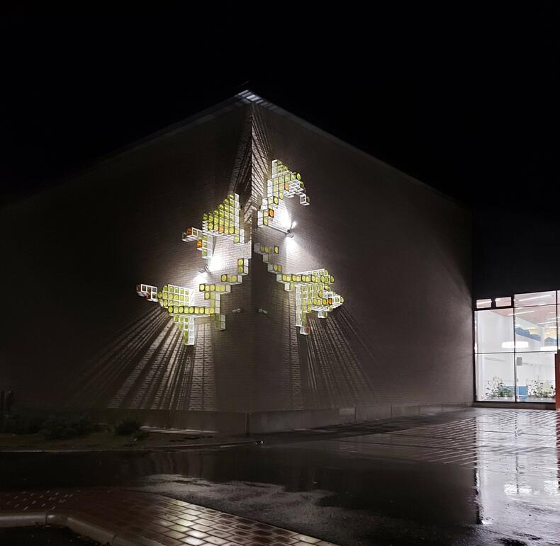 Julkinen taideteos Auringon kulma iltavalaistuksessa Luonnonmaan perhetalon ulkoseinässä.