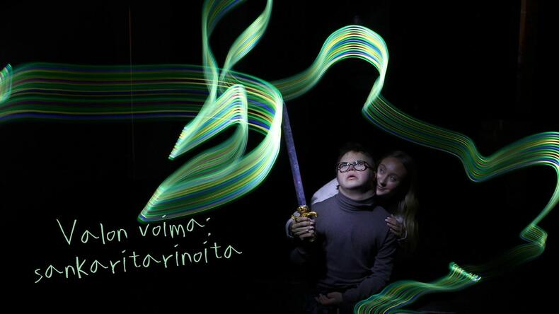 Mustalle taustalla valotaidetta tekevä poika pyörätuolissa avustaja vierellään. 