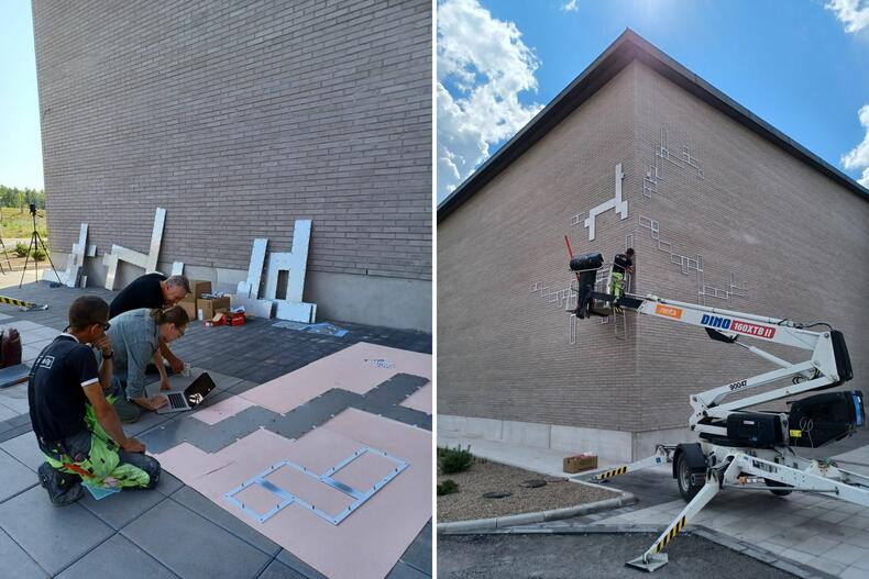 Vasemmalla työmiehet ja taiteilija katsovat maahan kumartuneina ruutua, oikealla nosturilla kiinnitetään levyjä rakennuksen julkisivuun.