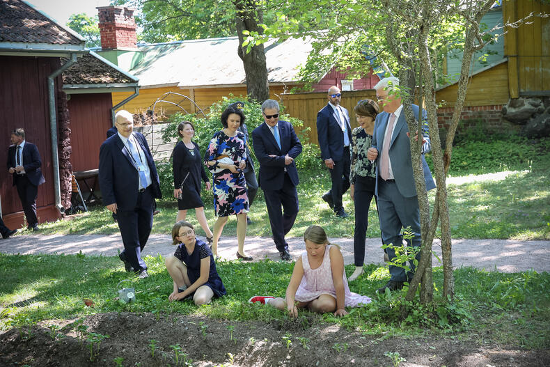 Presidenttejä vaimoineen puutarhassa, etualalla istuu kaksi tyttöä perunamaan reunalla