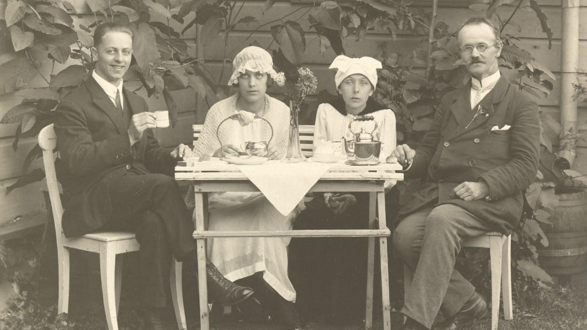 Mustavalkokuvassa istuu kaksi miestä ja kaksi naista kahvipöydän ympärillä