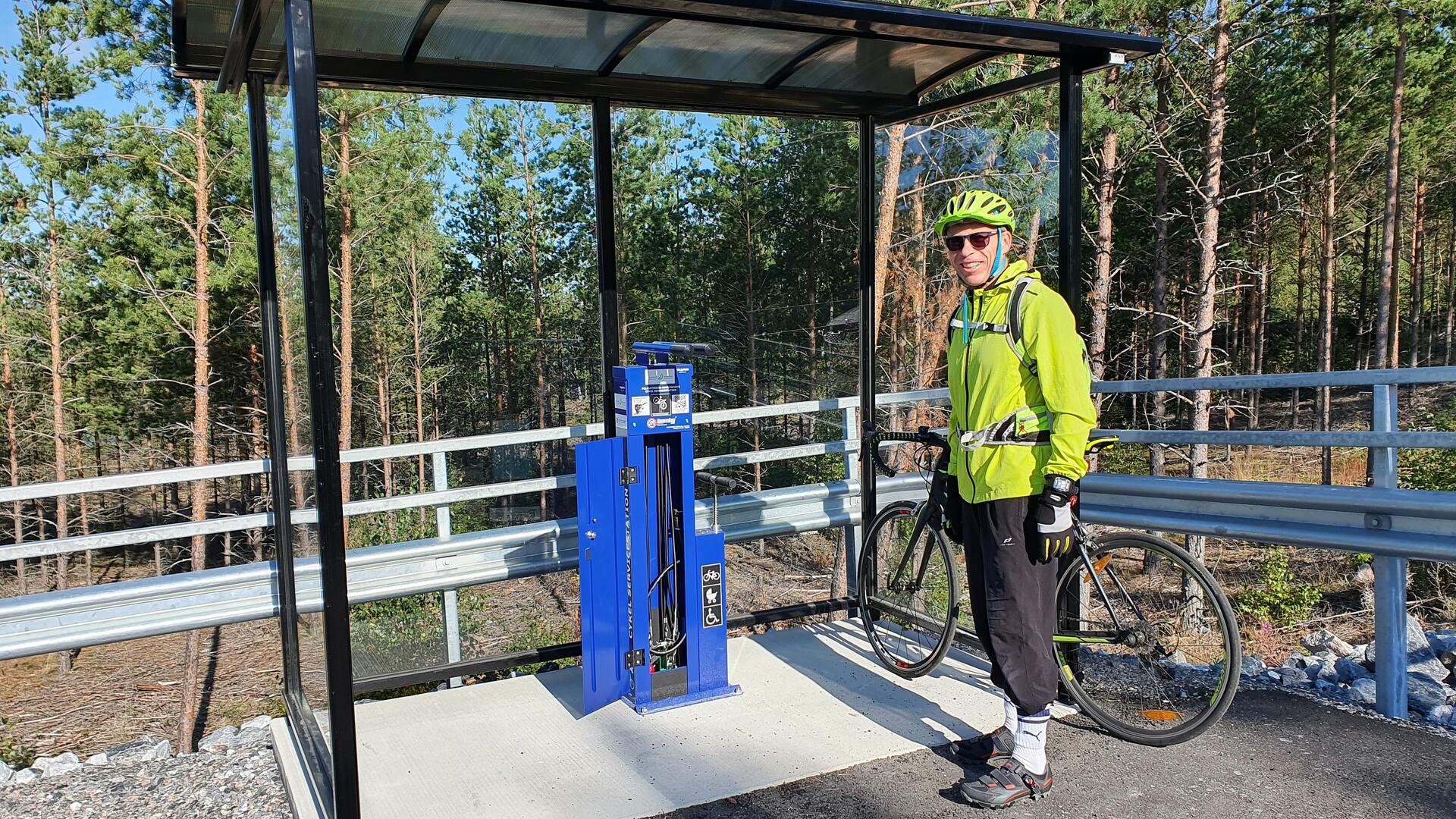 Vihreään kypärään ja urheilutakkiin pukeutunut pyöräilijä seisoo pyöränsä vieressä huoltopisteen edustalla. 