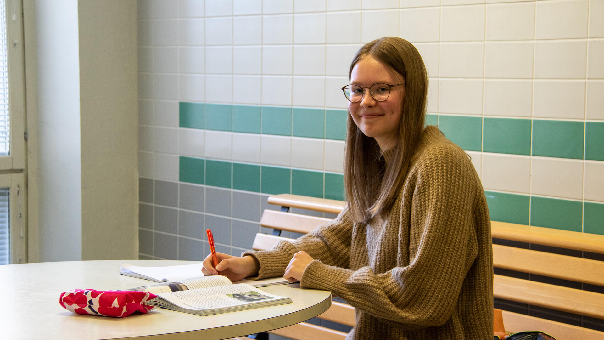 Nuori oppilas istuu pöydän ääressä tehden muistiinpanoja vihkoon, kirja vieressä.