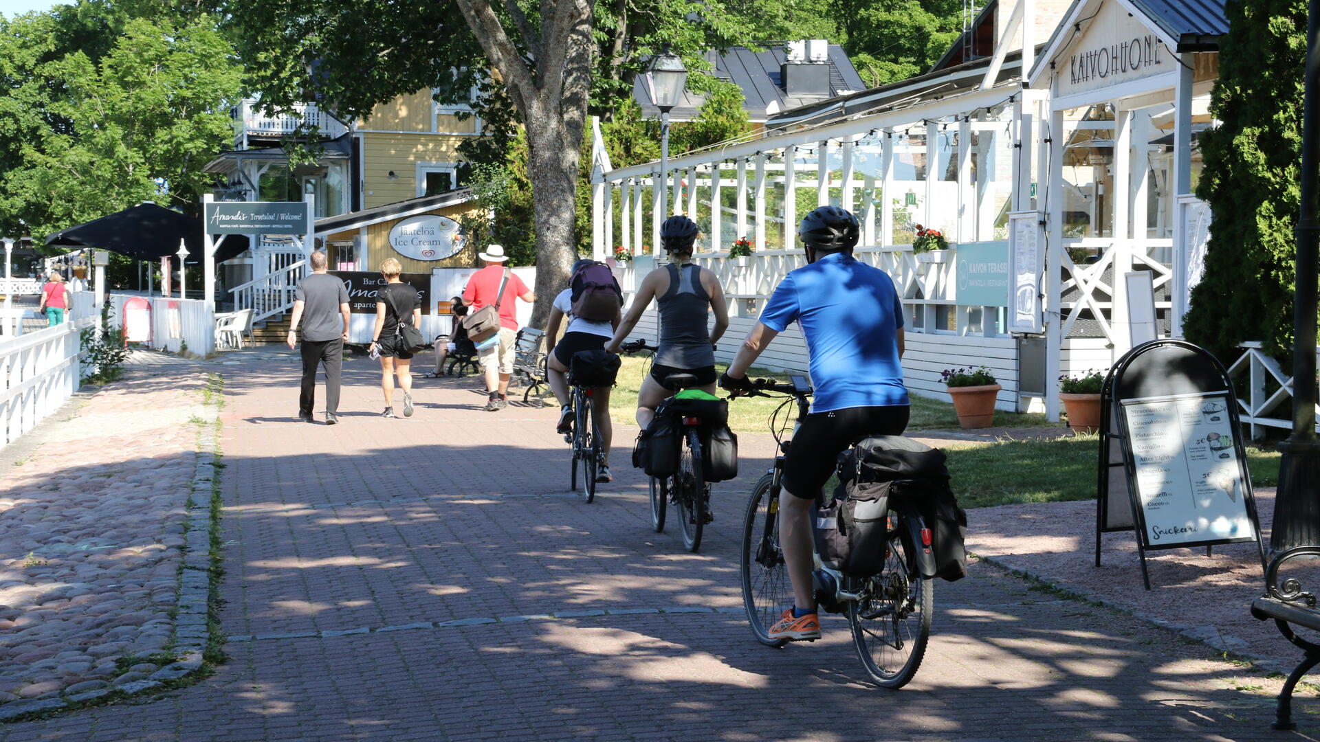 Kolme pyöräilijää ajaa Vanhassakaupungissa ravintola Kaivohuoneen edustalla, kesäisenä päivänä rannassa liikkuu ihmisiä.