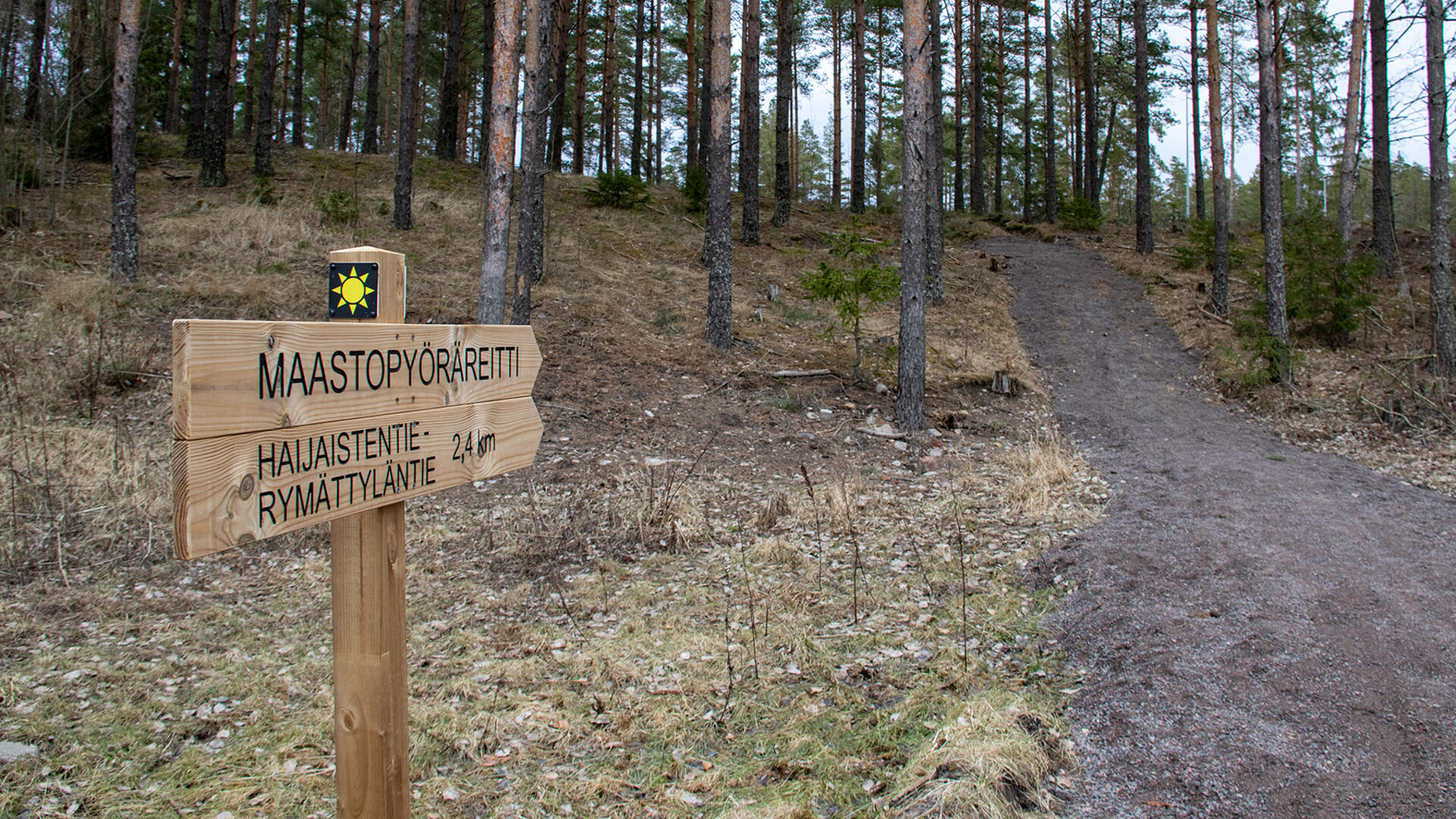 Hiekkatie vie ylämäkeen metsään, tien vieressä on kyltti, joka osoittaa ylämäkeen: Maastopyöräreitti, Haijaistentie-Rymättyläntie 2,4 km.