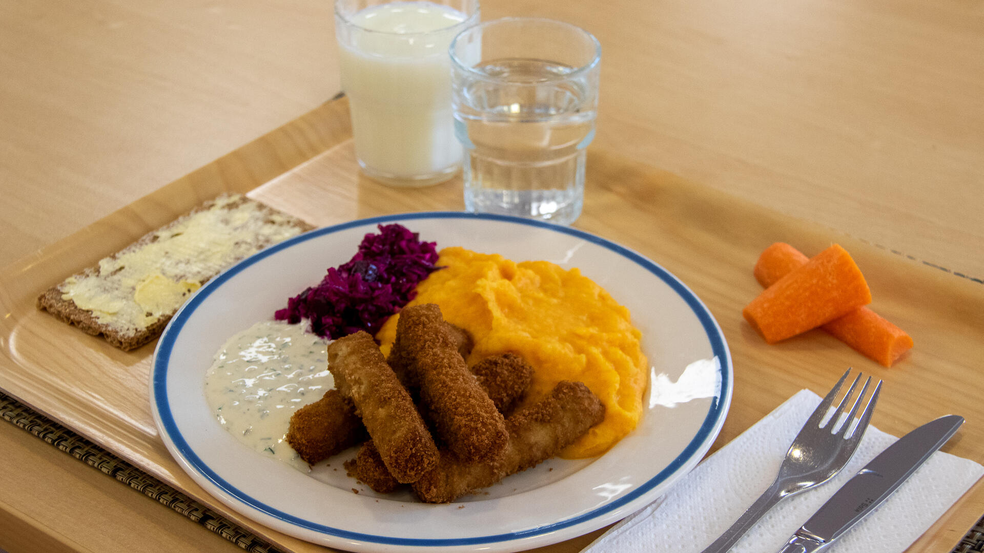 Kouluruokatarjottimella on lasi vettä ja maitoa, aterimet servetillä, voideltu näkkäri, kolme porkkanapalaa keossa sekä keskellä lautanen, jolle on koottu juuresmuusia, viisi kalapuikkoa kasaan sekä kermaviilikastiketta ja punajuurenväristä raastesalaattia.