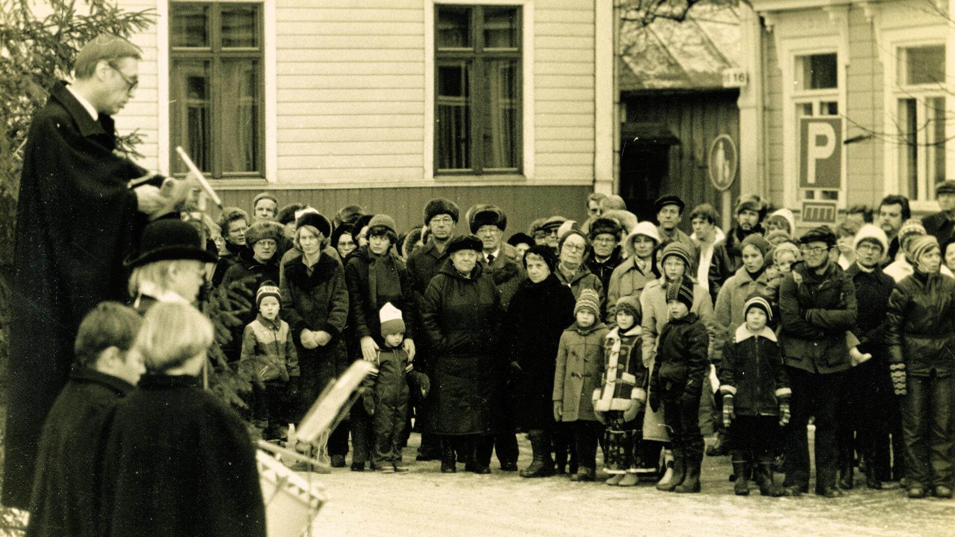 mustavalkoinen kuva, jossa joulurauhaa julkistava mies seisoo korokkeella vasemmassa reunassa ja yleisö jonkin matkan päässä hänestä kuvan taustalla ja oikeassa reunassa. Takana puurakennuksia ja puhujan takana häämöttää joulukuusen oksia.