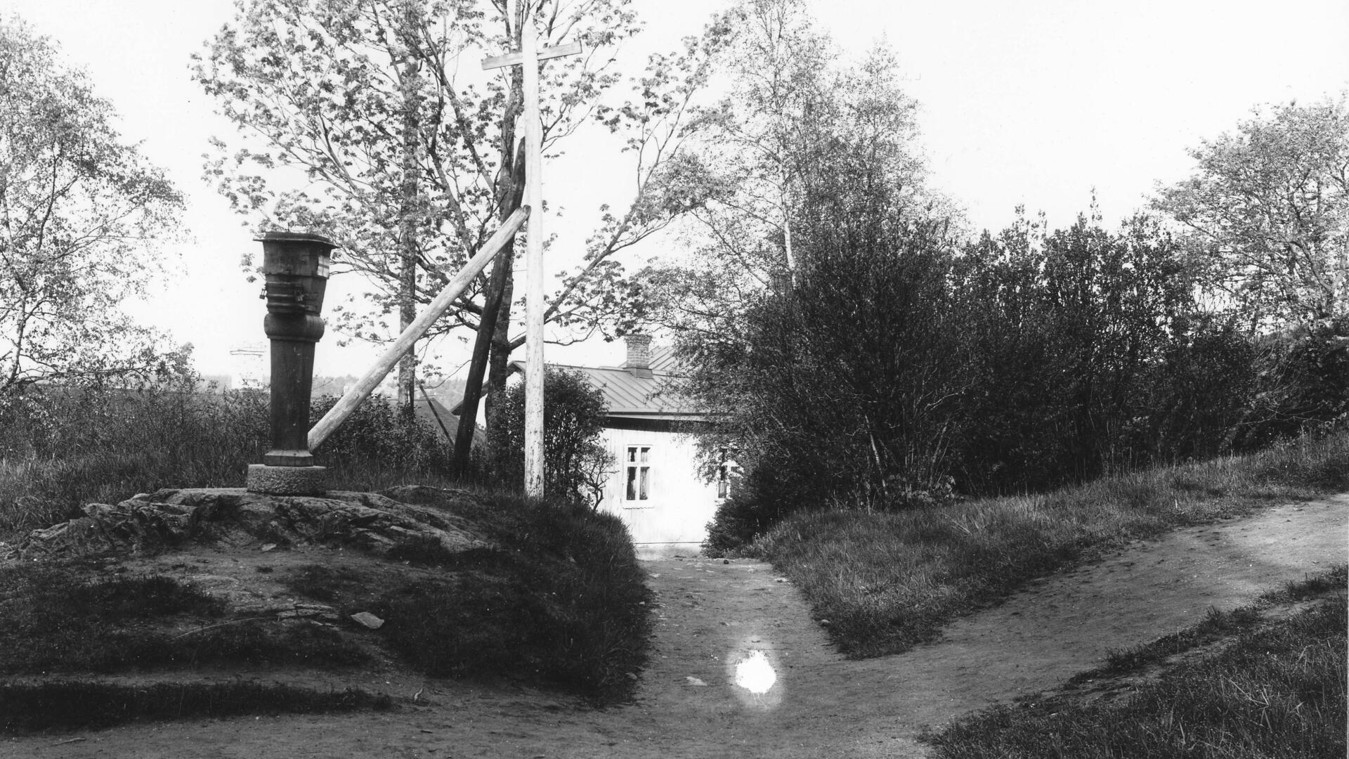 ulkokuva, jossa näkyy soraiset kävelytiet ja vasemmalla olevalla kummulla köyhäintukki. Taustalla näkyy osa vanhasta puurakennuksesta. 