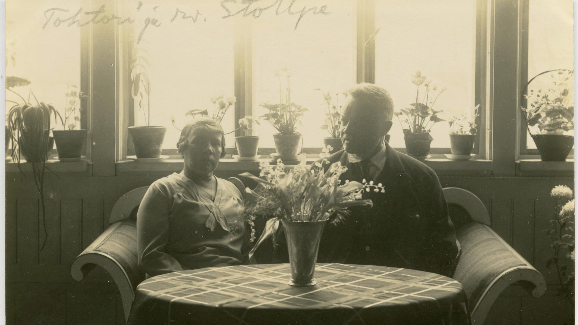 Mustavalkoinen vanha valokuva, jossa pariskunta istuu ikkunan edessä puisella sohvalla, edessään pöytä.