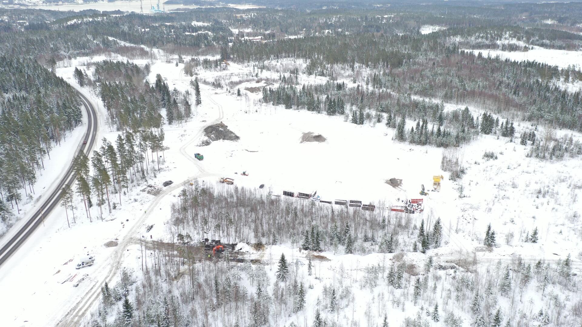 Talvinen maisema ilmasta kuvattuna: vasemmalla kulkee tie, oikealla näkyy paljon metsää ja keskellä raivattua maata, jossa liikkuu työkoneita.