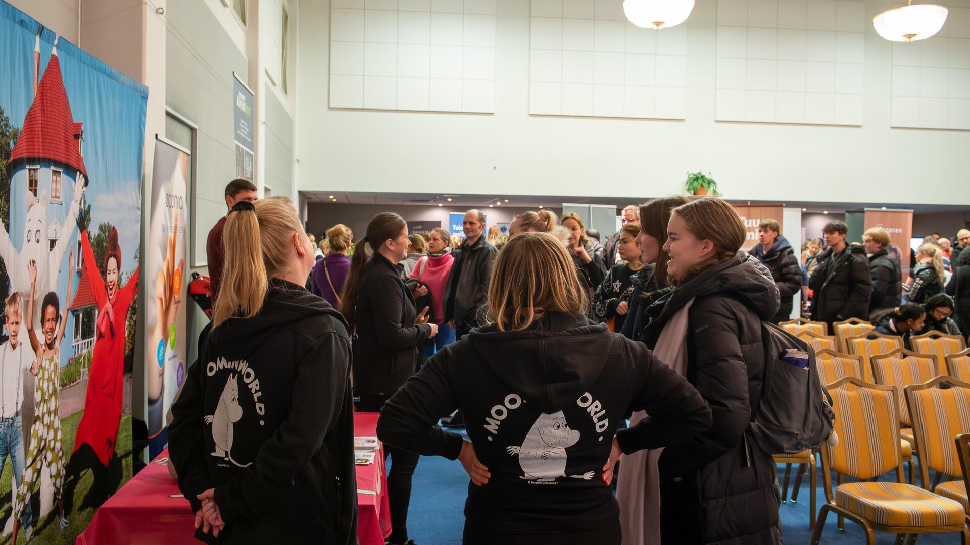 Muumihuppareihin pukeutunutta henkilökuntaa keskustelemassa messuvieraiden kanssa tapahtumassa.