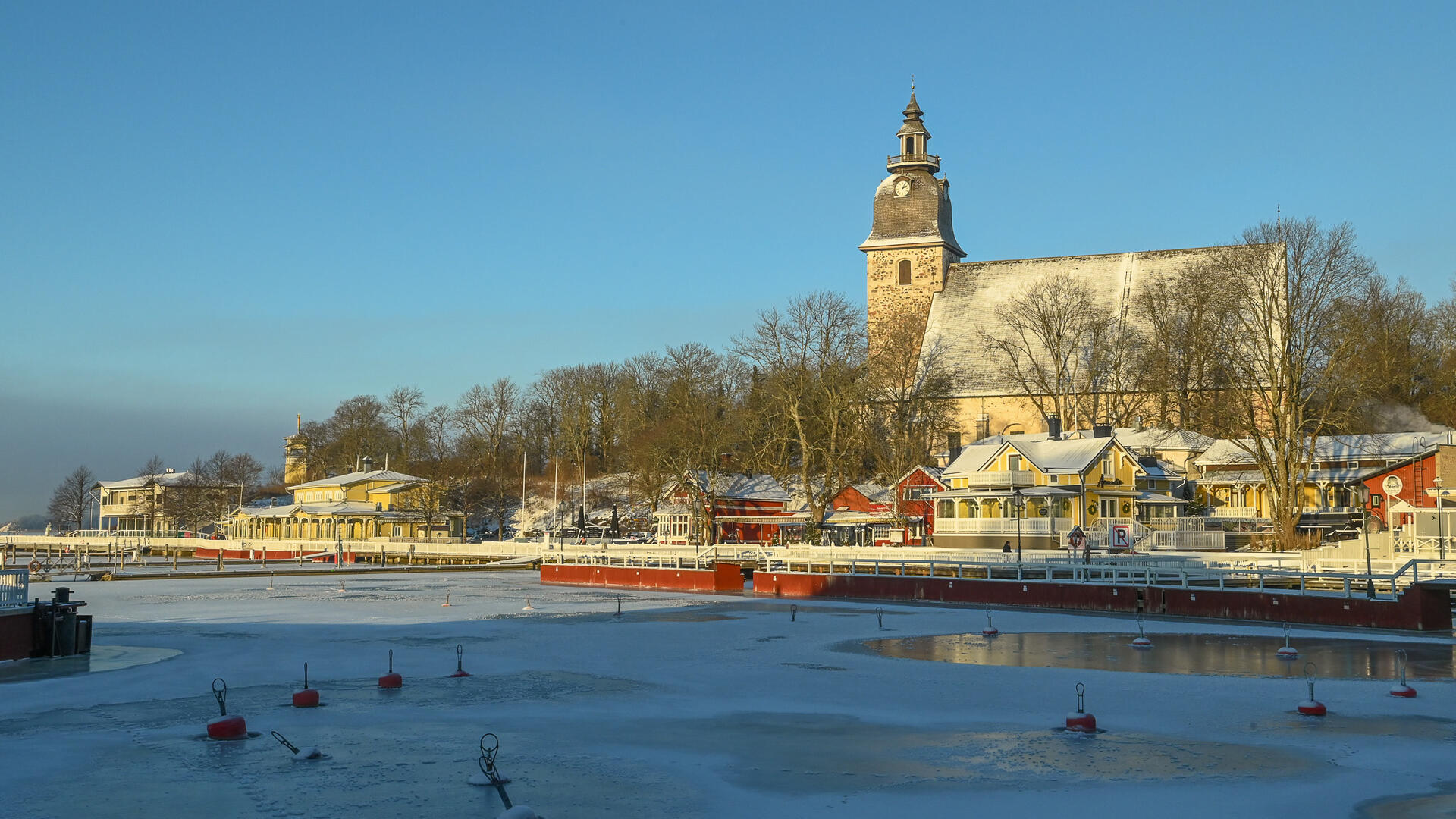 Naantalin vierasvenesatama talvella, rannan kiinteistöjen takana auringonpaisteessa Naantalin kirkko.