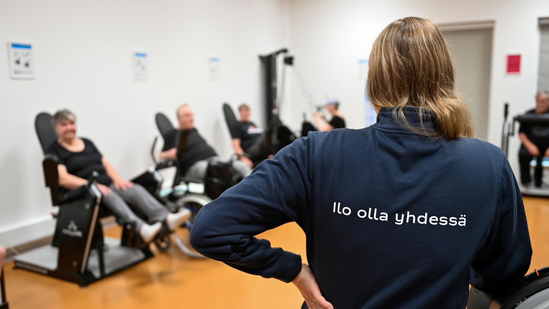 Soveltavan liikunnan asiakkaita kuntosalilla, etualalla ohjaaja, jonka selässä lukee Ilo olla yhdessä.