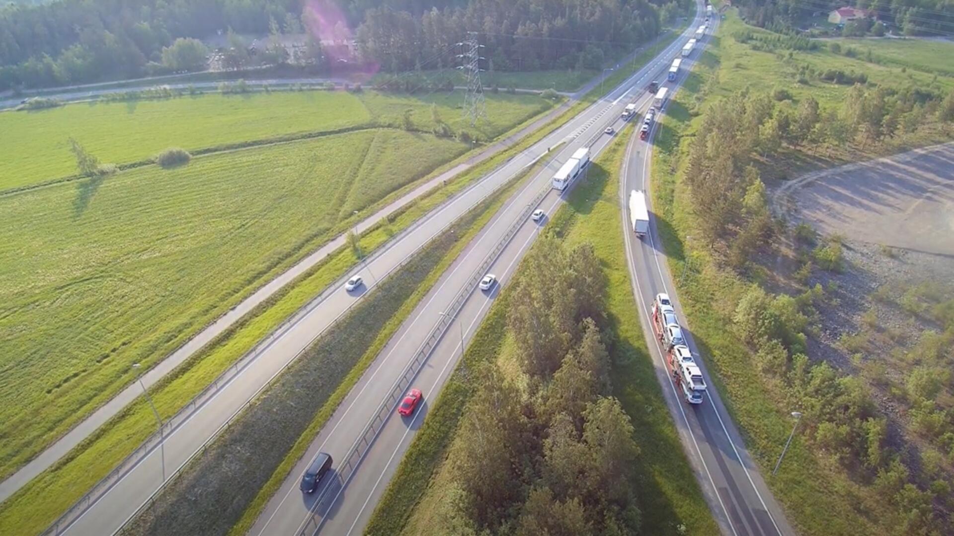 Ylhäältä kuvattuna näkyy E18-tie, jossa rekat ja autot ajavat. Kaistoja liittyy tiehen, ympärillä on vehreää pelto- ja metsämaisemaa.