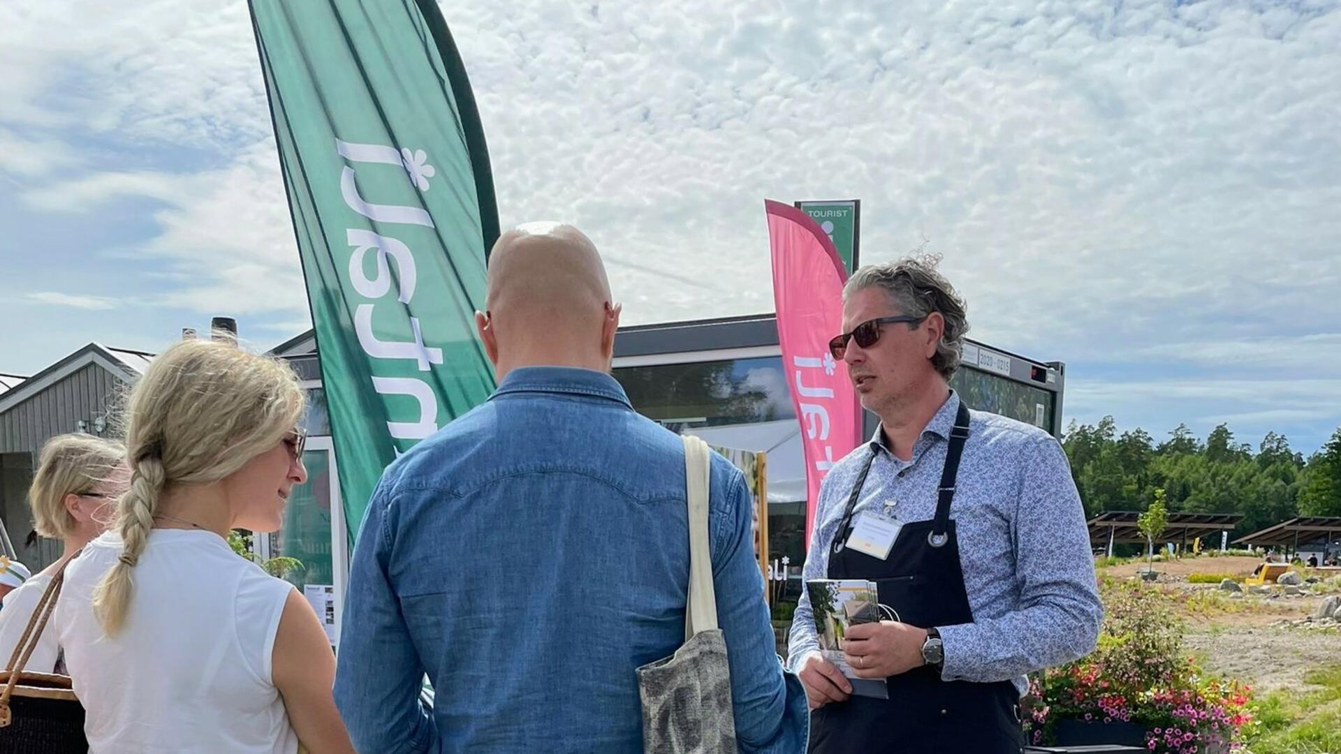 Mustaan essuun pukeutunut mies juttelee ihmisten kanssa, taustalla Naantali-lippuja.