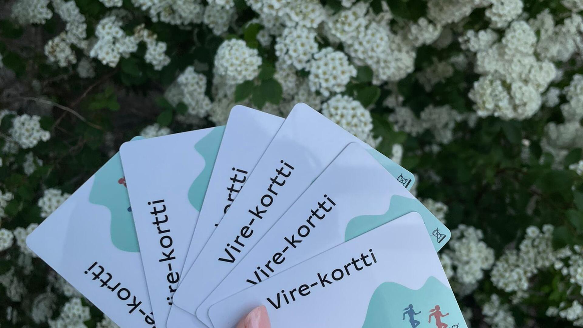 Kuvassa taustakuvana valkoiset kukat, sekä käsi, jossa useita virekortteja.