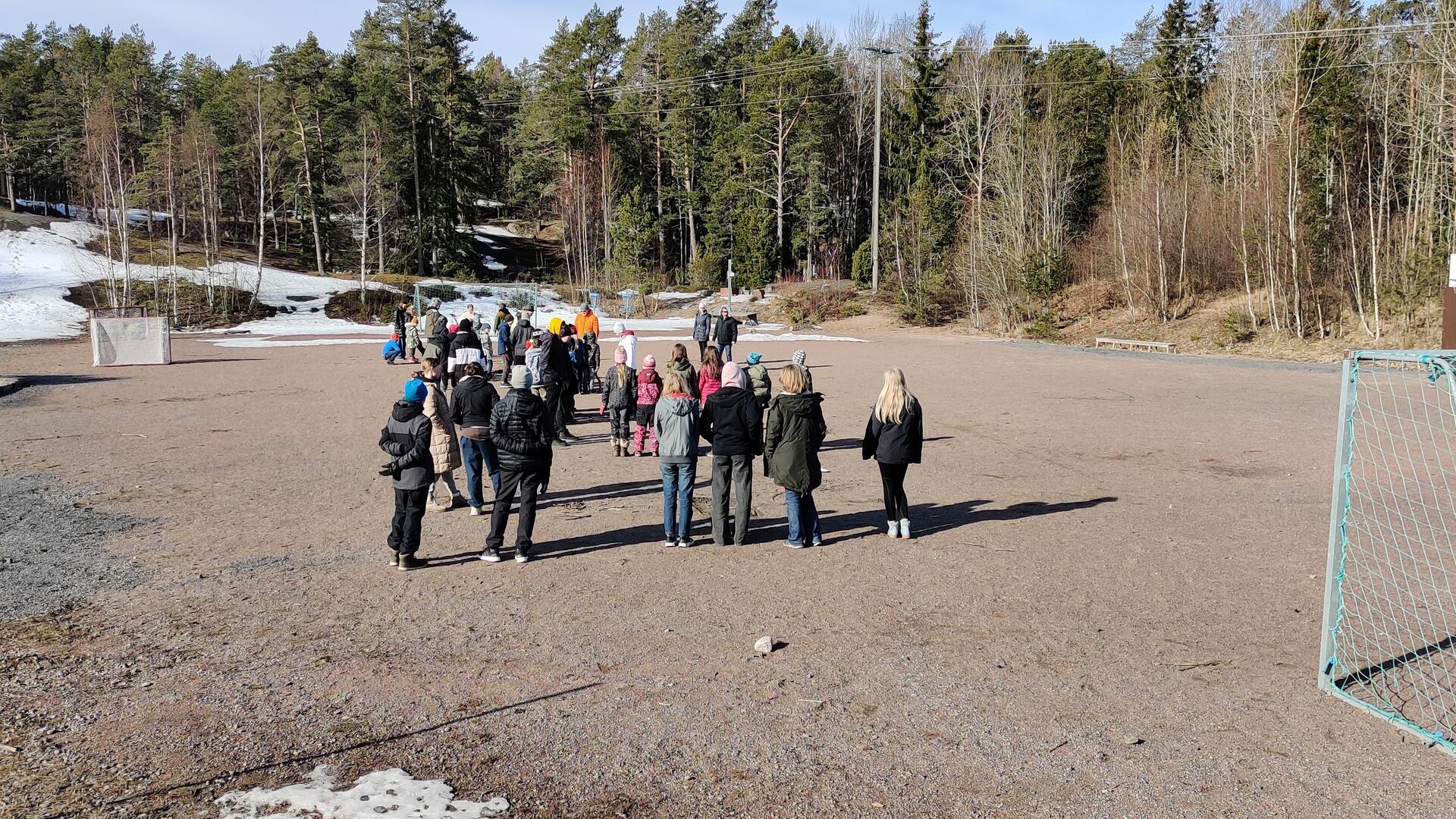 Oppilaita seisoo heikkakentällä koulun pihalla kuuntelemassa aikuisia.