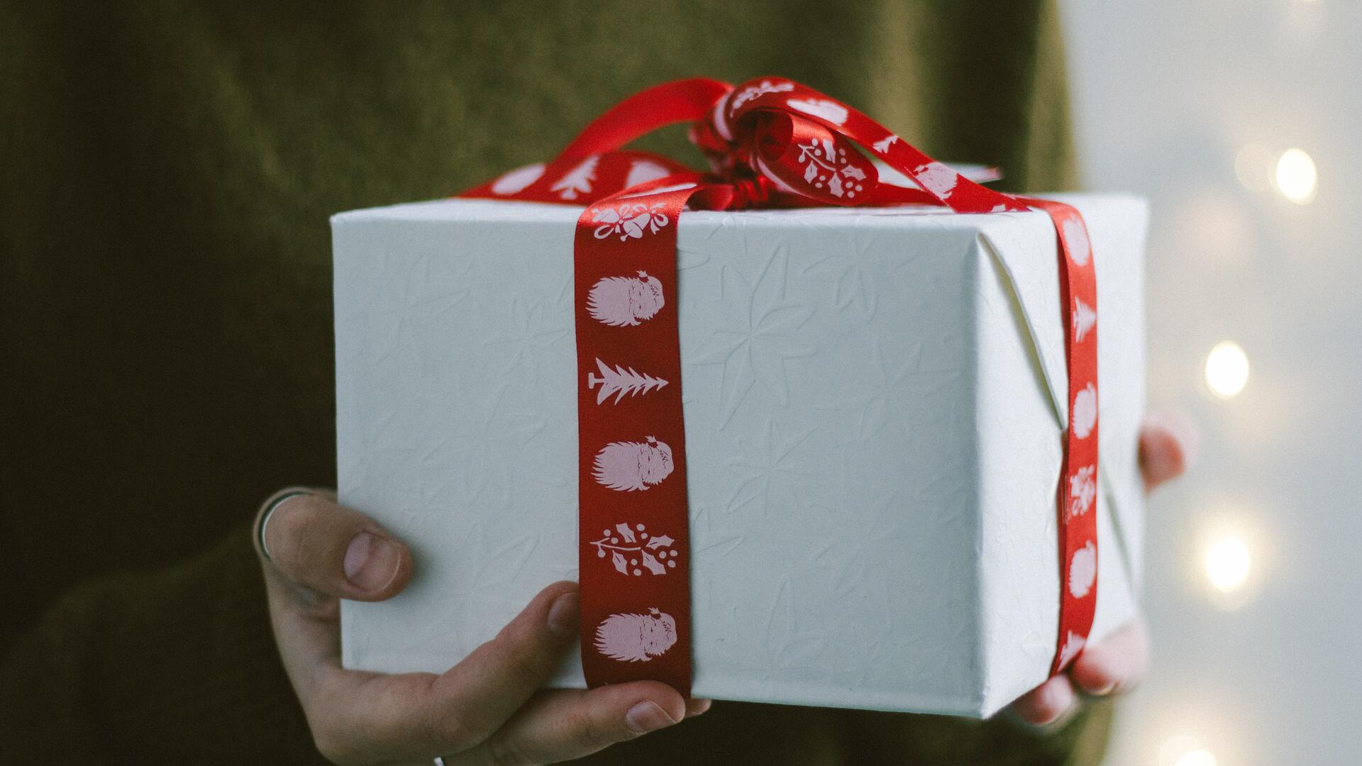 Henkilö pitelee käsissään kuution muotoista valkoiseen paperiin paketoitua lahjapakettia, jossa on punainen joulunauha.