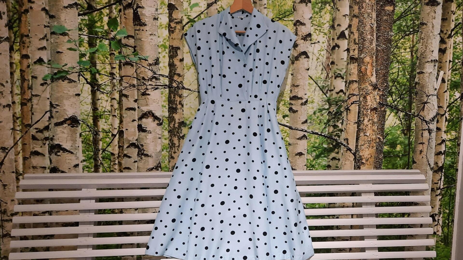Taustalla kuva koivumetsästä ja kuvan edessä valkoinen penkki, jonka päällä vaaleansininen kesämekko, jossa on mustia pilkkuja. 