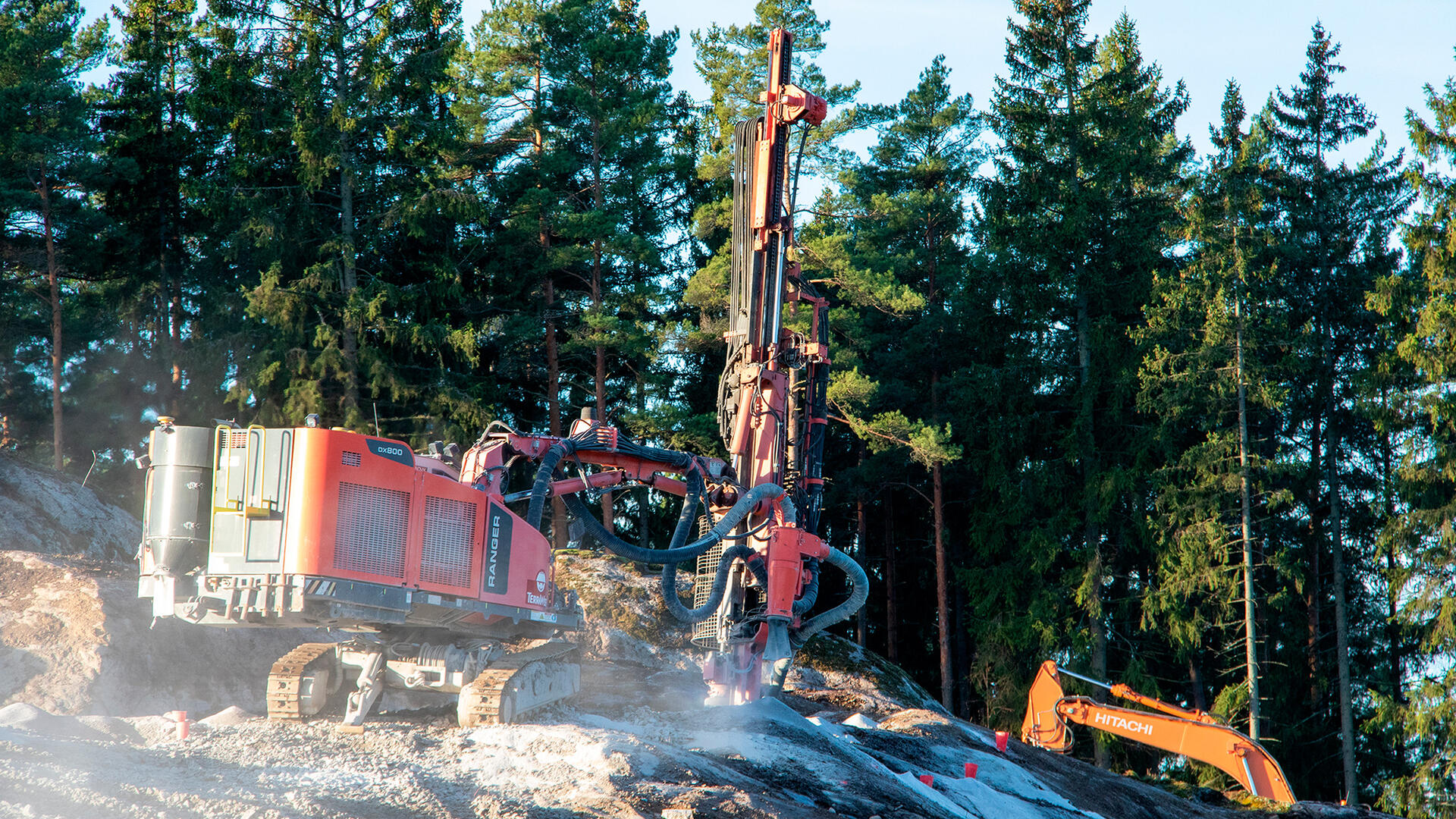 Työmaakone murskaa kalliota, taustalla kohoavat kuusipuut. Taaempana työn touhussa on myös kaivinkone.