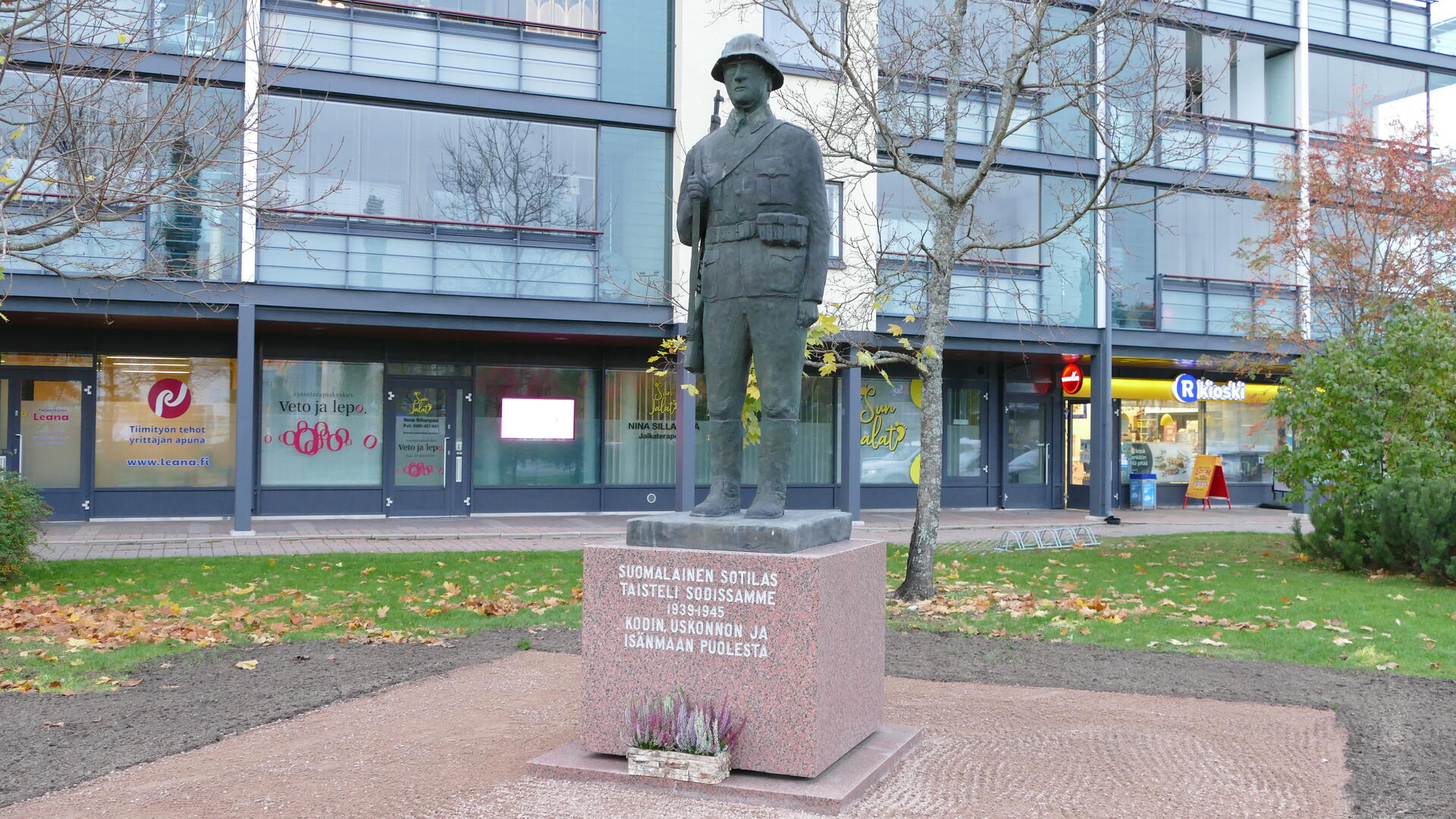 keskellä sotilaspatsas kivipaadella, ympärillä puistomaisemaa ja taustalla kerrostalo, jonka alakerrassa liiketiloja