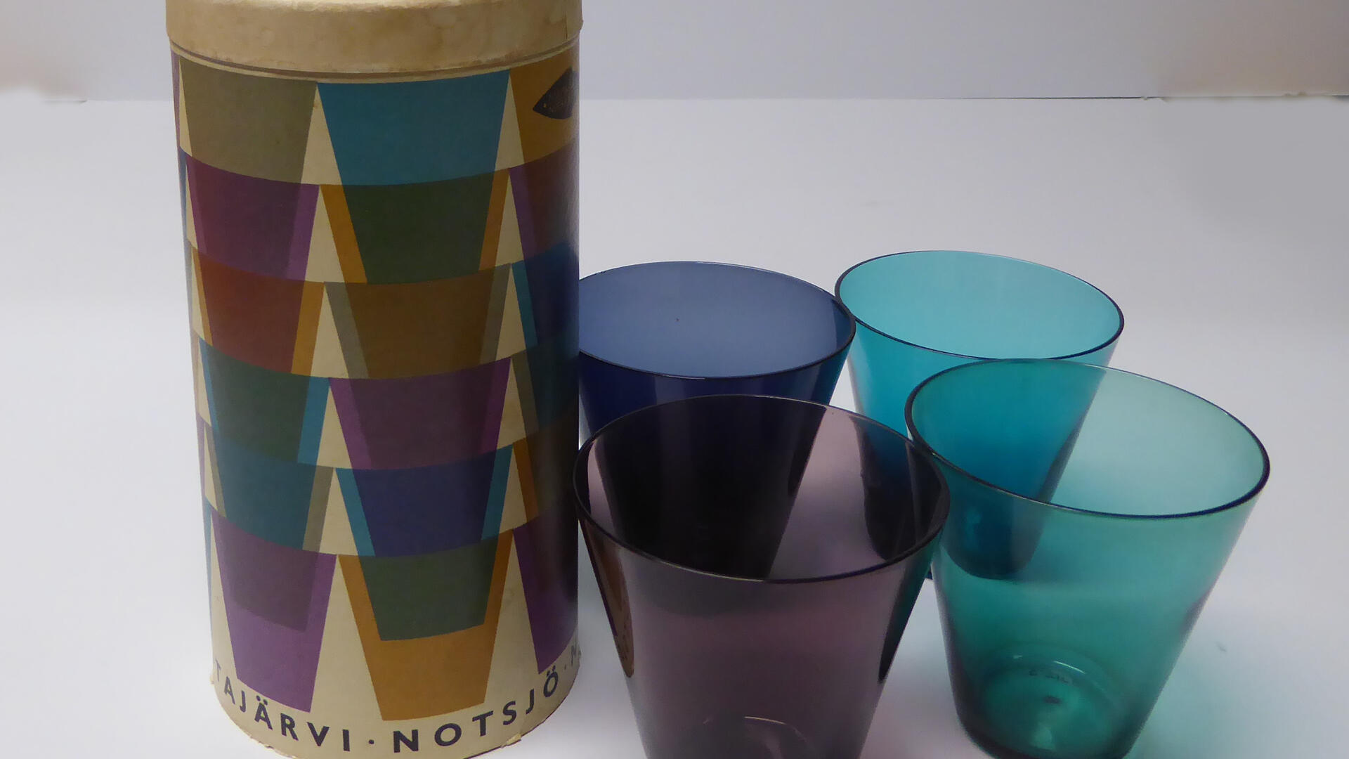 Neljä eriväristä juomalasia ja niiden pyöreä, pahvista tehty myyntipakkaus