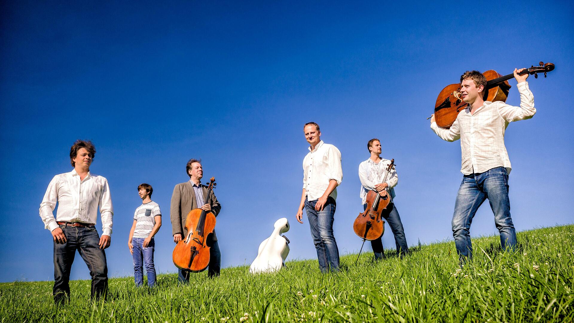 Vihreällä ruoholla sinistä taivasta vasten on useampi farkkuihin ja valkoisiin paitoihin pukutunut mies sellojen kanssa.