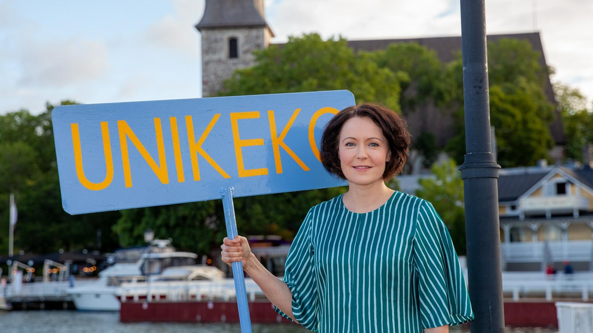 Jenni Haukio raitaan pukeutuneena Naantalin Vanhankaupungin rannassa kädessän Unikeko-kyltti.