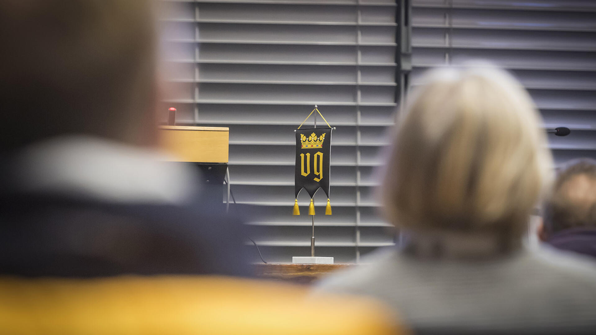 Naantalin musta-keltainen vaakuna pöytäviirissä pöydällä valtuustosalissa, etualalla ihmisiä.