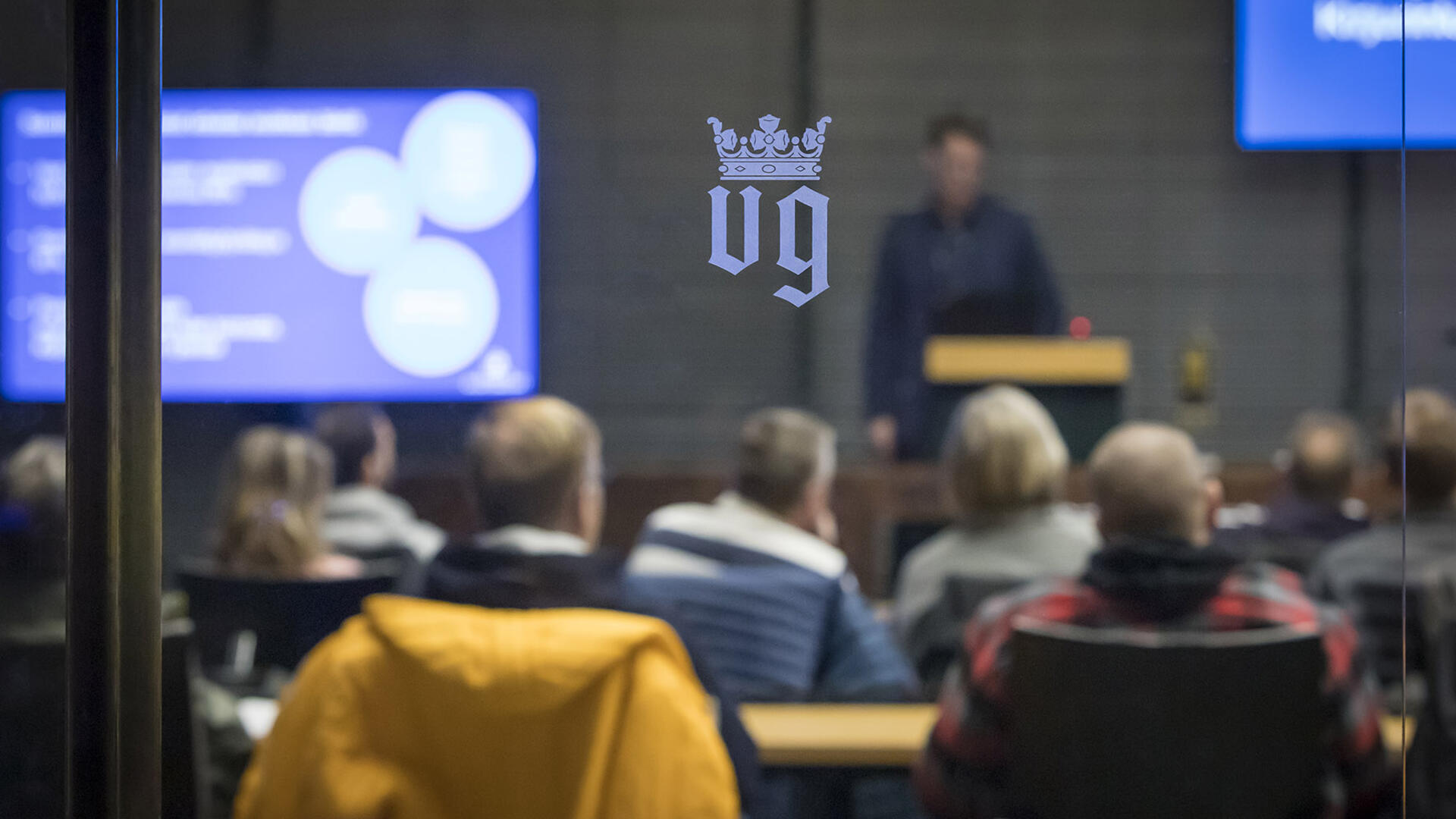 Naantalin VG-vaakuna lasiovessa, oven takana käynnissä yleisötilaisuus kaupungintalon valtuustosalissa.