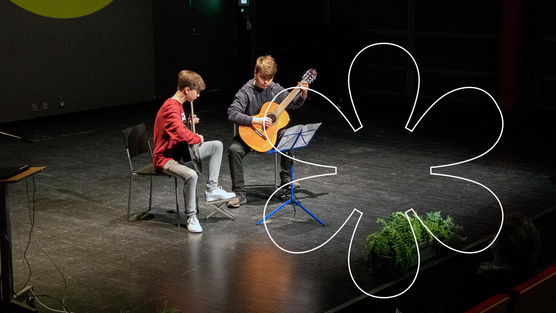 Kaksi nuorta poikaa istuu vierekkäin valokeilassa lavalla kitaraa soittaen, edessään nuottiteline. Kuvan päällä Onnellinen aurinko -elementti.