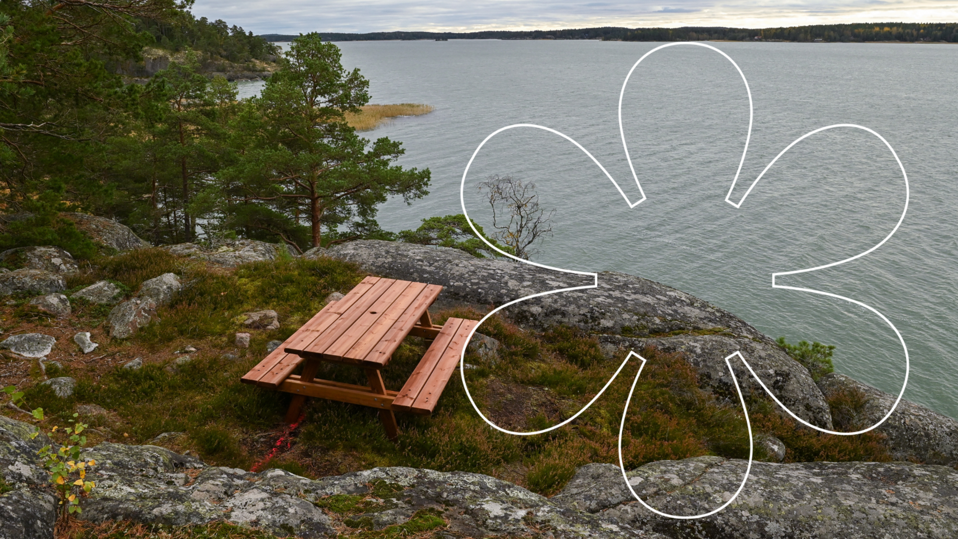 Pöytäryhmä kallioilla, josta avautuu maisema merelle. Onnellinen aurinko -tunnus.