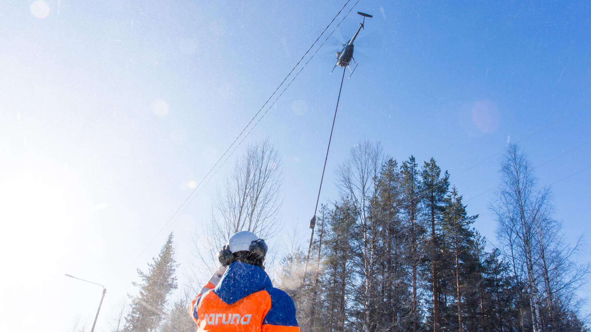 Carunan työntekijä seisoo työmaakypärä, kuulosuojaimet ja oranssi takki päällä metsän edustalla, sähkölinjojen yläpuolella on helikopteri.