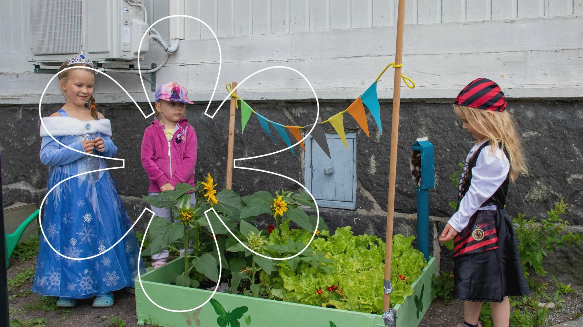 Kolme lasta istutuslavan ympärille kerääntyneenä, laatikossa on pieni kasvimaa ja se on koristeltu mm. värikkäin viirein.
