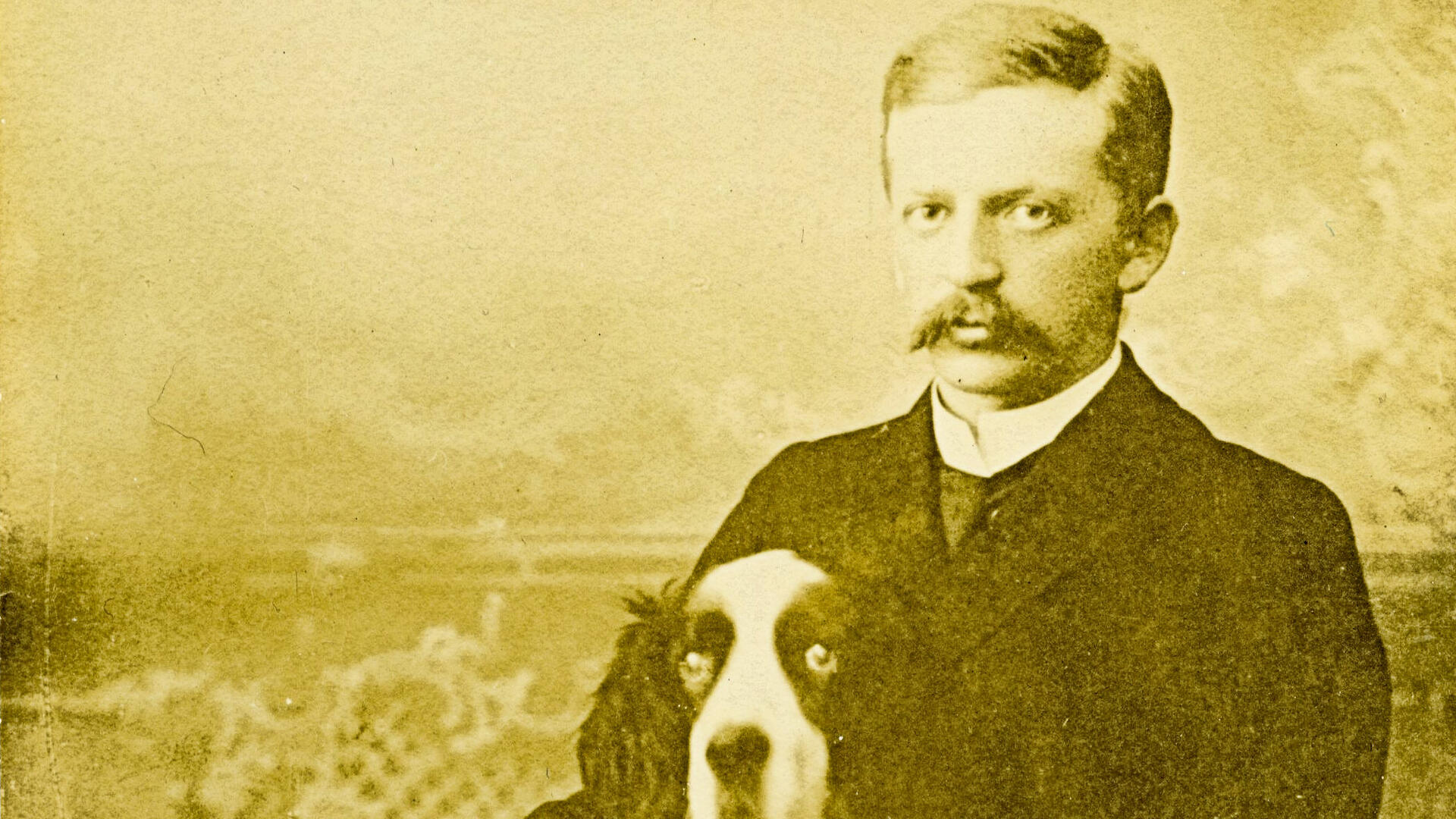 Vanhassa mustavalkokuvassa mies istuu tuolissa ja hänen edessään on koira, joka on puoliksi miehen sylissä