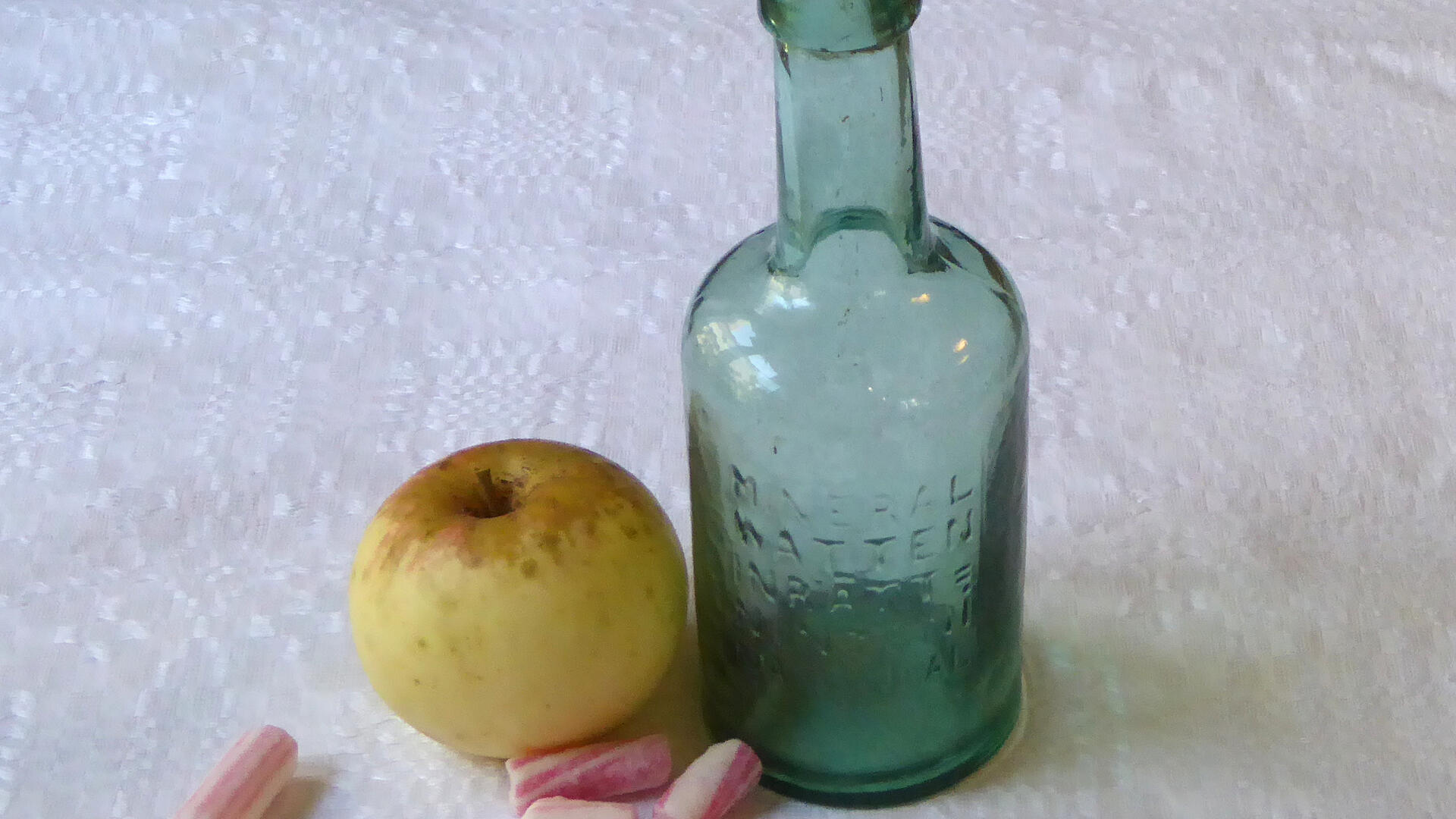valkoisella taustalla vihreä lasipullo, omena ja karamelleja